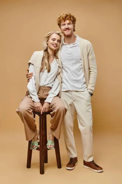 Full length of redhead man embracing blonde woman sitting on chair and looking at camera on beige — Stock Photo