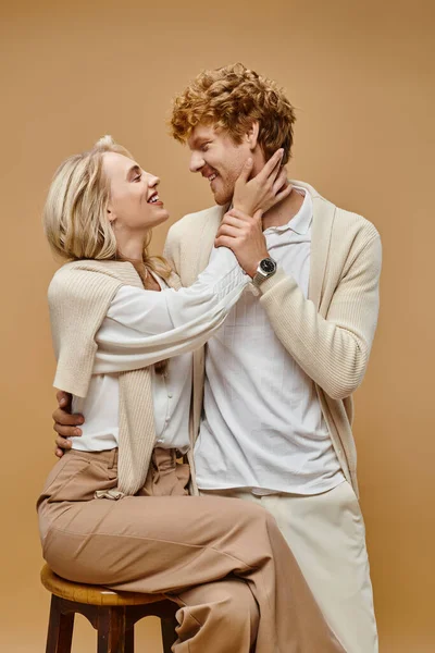 Mulher loira feliz abraçando homem ruiva na moda enquanto sentado na cadeira em bege, moda estilo clássico — Fotografia de Stock