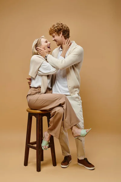 Laughing blonde woman sitting on chair and hugging young redhead man on beige, old money style — Stock Photo