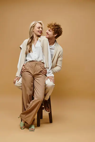 Homme rousse joyeux assis sur une chaise près de femme blonde à la mode sur beige, mode classique intemporelle — Photo de stock