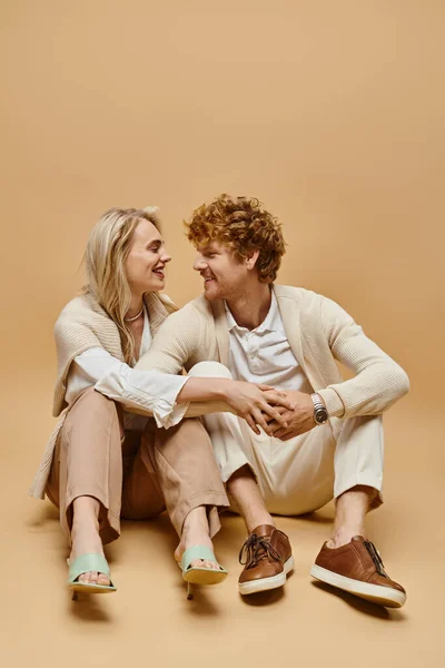Cheerful young couple in light-colored clothes sitting and smiling at each other on beige backdrop — Stock Photo