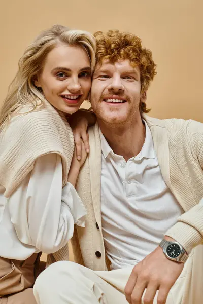 Pleased blonde woman and redhead man in light-colored outfit looking at camera on beige backdrop — Stock Photo