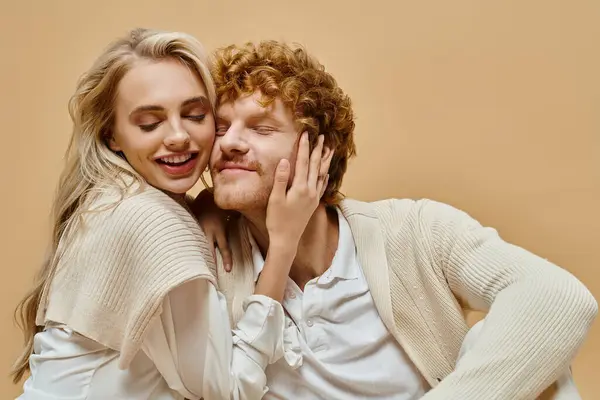 Mulher loira encantada com olhos fechados abraçando cabeça de homem ruiva elegante no fundo bege — Fotografia de Stock