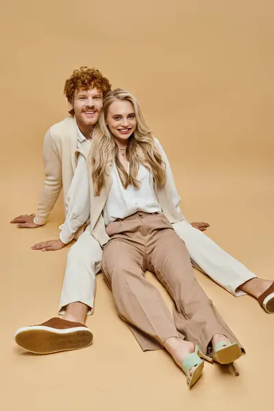 Carefree stylish couple in light-colored clothes sitting and looking at camera on beige backdrop — Stock Photo