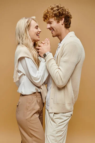 Vista lateral do casal na moda roupas de cor clara de mãos dadas e rindo em pano de fundo bege — Fotografia de Stock