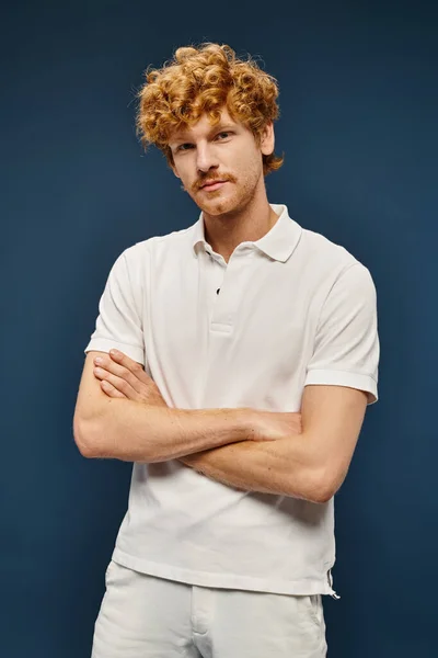 Homme rousse à la mode en polo-shirt blanc regardant caméra avec bras croisés sur bleu, mode classique — Photo de stock