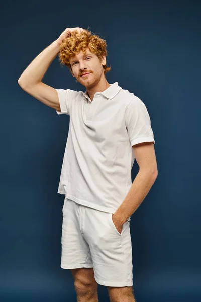 Рудий чоловік у білій футболці і шортах, що стоять з паличкою в кишені на синьому, позачасовому моді — стокове фото