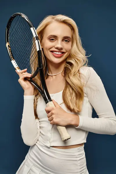 Радісна спортивна блондинка в білому вбранні тримає тенісний ракетка і посміхається на камеру на синьому — стокове фото