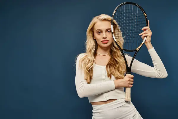 Trendy wealthy blonde woman in white outfit with tennis racquet looking at camera on blue backdrop — Stock Photo