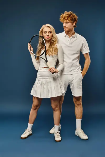 Full length of redhead man near blonde woman with tennis racquet on blue, aristocratic couple — Stock Photo