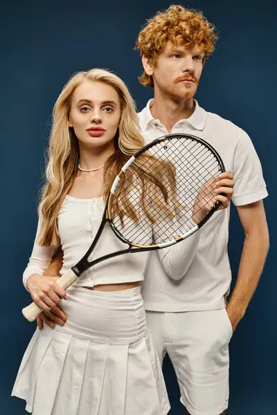 Stylish blonde woman with tennis racquet looking at camera near redhead man in white clothes on blue — Stock Photo