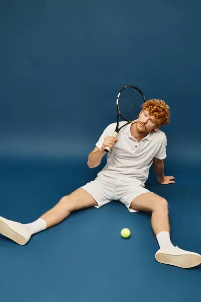 Homem ruivo esportivo em roupas brancas da moda com raquete sentado perto de bola de tênis no fundo azul — Fotografia de Stock