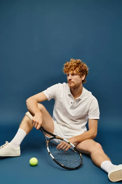 Homme rousse à la mode en vêtements de tennis blanc avec raquette assis près de la balle de tennis sur fond bleu — Photo de stock