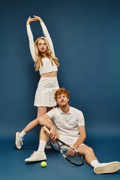 Trendy blonde woman in white tennis outfit with raised hands near redhead man with racquet and ball — Stock Photo