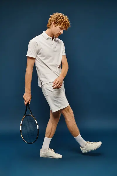 Повна довжина молодого рудого чоловіка в білому модному вбранні, що йде з тенісним ракеткою на синьому фоні — стокове фото