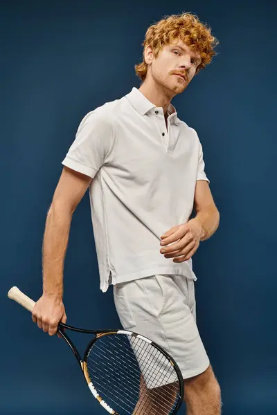 Добре одягнений рудий чоловік в білому тенісному вбранні тримає ракетку і дивиться на камеру на синьому — стокове фото