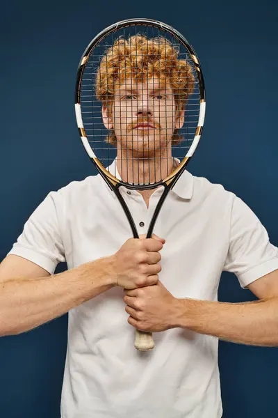 Trendy redhead man in white clothes obscuring face with tennis racquet and looking at camera on blue — Stock Photo