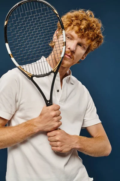 Guapo pelirrojo en traje de tenis blanco sosteniendo raqueta cerca de la cara en azul, moda atemporal - foto de stock