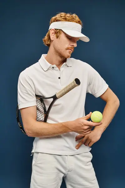 Jovem ruivo homem em roupas brancas e viseira sol segurando raquete e olhando para bola de tênis em azul — Fotografia de Stock