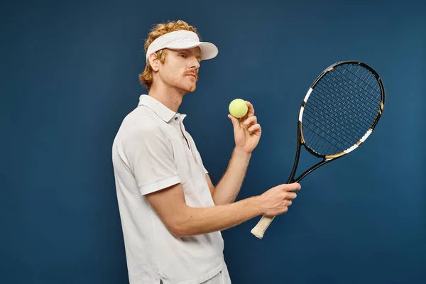 Giovane bel rossa uomo in abiti bianchi e visiera solare con racchetta e palla da tennis su blu — Foto stock