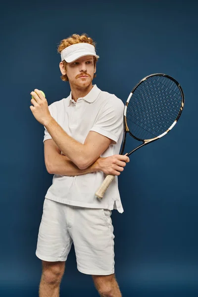 Rossa uomo in abito bianco e visiera solare in possesso di racchetta e palla da tennis, mentre guardando altrove su blu — Foto stock