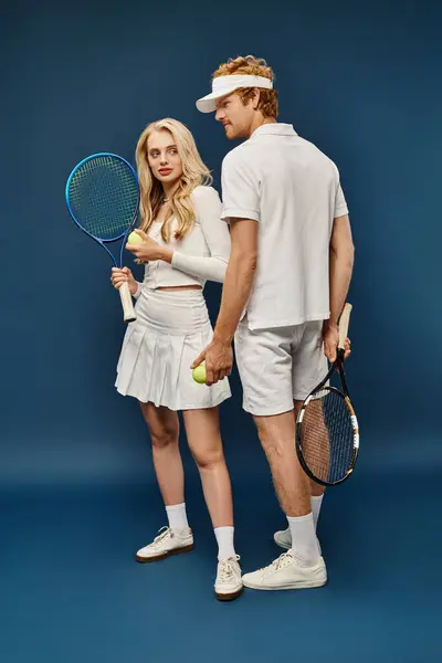 Full length of young couple in white tennis attire with racquets and balls on blue, old money style — Stock Photo