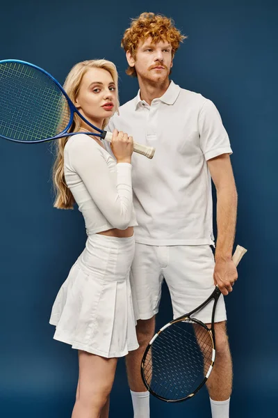Old money style, young trendy couple in white tennis outfit posing with racquets on blue backdrop — Stock Photo