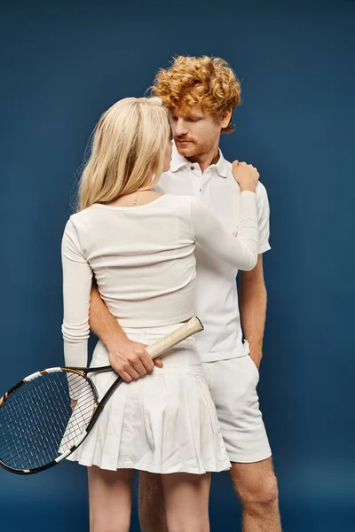 Mujer rubia de moda abrazando joven pelirrojo en ropa blanca con raqueta de tenis en azul - foto de stock