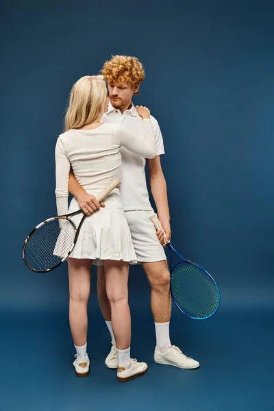 Повна довжина блондинки, обіймаючи молодого рудого чоловіка в білому одязі з тенісним ракеткою на синьому — стокове фото