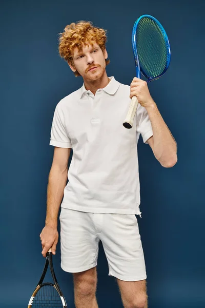Homem ruivo bonito em traje branco com raquetes de tênis olhando para longe em azul, estilo de vida ativo — Fotografia de Stock