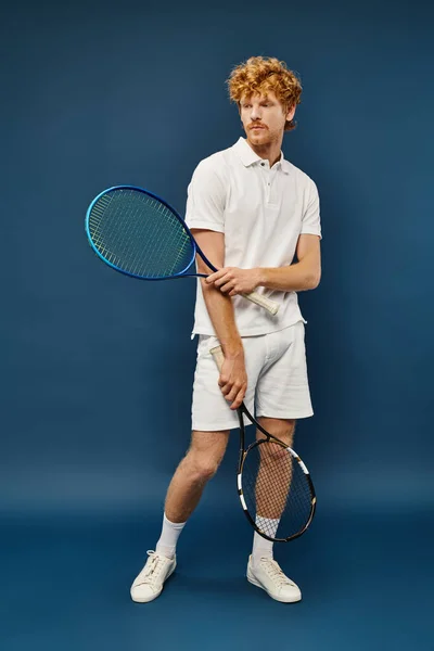 Comprimento total do jovem ruivo em traje branco com raquetes de tênis olhando para o fundo azul — Fotografia de Stock