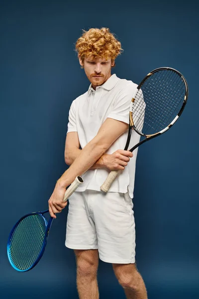 Aristocrático pelirrojo en traje blanco posando con raquetas de tenis posando sobre fondo azul - foto de stock