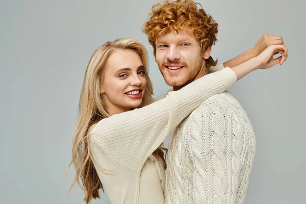 Cheerful blonde woman embracing young happy redhead man on grey, trendy old money style couple — Stock Photo
