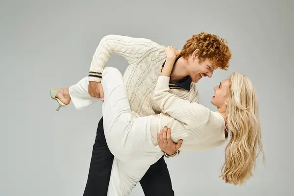 Verspielter rothaariger Mann in lässiger Kleidung umarmt trendige junge blonde Frau vor grauem Hintergrund — Stockfoto