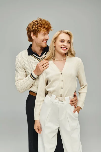 Feliz ruiva homem em traje casual abraçando jovem loira no fundo cinza, casal na moda — Fotografia de Stock
