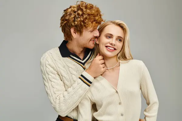 Playful redhead man touching neck of joyful blonde woman in trendy clothes on grey, classic fashion — Stock Photo