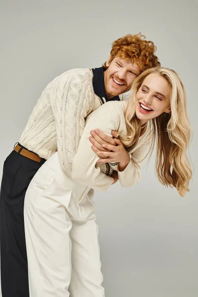 Joyful redhead man embracing laughing blonde woman and having fun on grey backdrop, classic fashion — Stock Photo