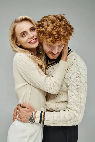 Lächelnde und stylische blonde Frau umarmt jungen rothaarigen Mann auf grauer, modischer Paarbeziehung — Stockfoto