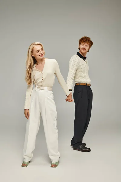 Full length of happy young redhead man and blonde woman holding hands on grey backdrop in studio — Stock Photo