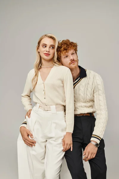 Dreamy redhead man hugging trendy blonde woman while sitting on white cube on grey, classic fashion — Stock Photo