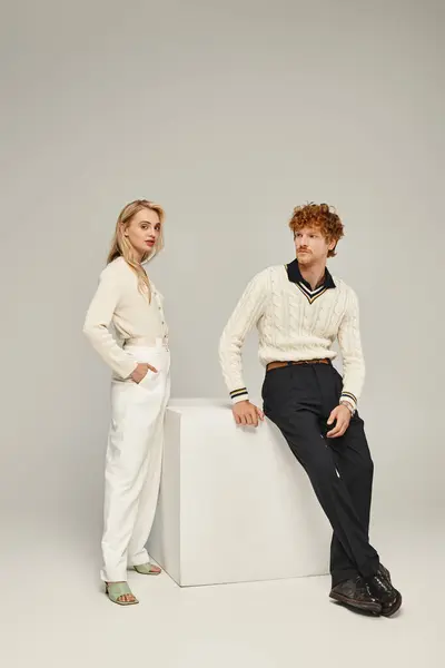 Blonde woman and redhead man in trendy casual attire posing near white cube on grey backdrop — Stock Photo