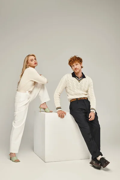 Rubia mujer y pelirroja hombre en elegante ropa casual posando cerca de blanco cubo en gris telón de fondo - foto de stock