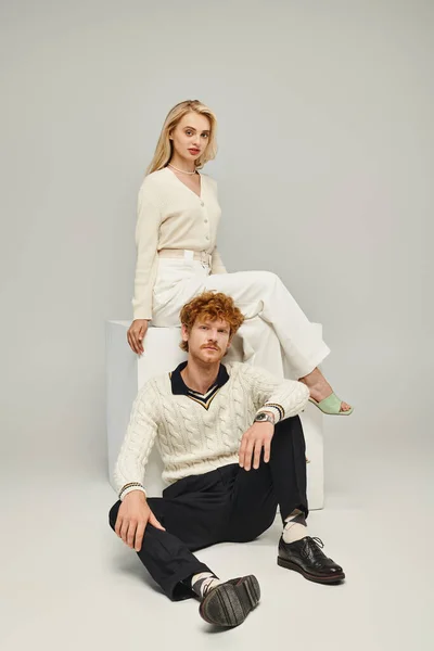 Trendy redhead man looking at camera near blonde woman sitting on white cube on grey, classic style — Stock Photo