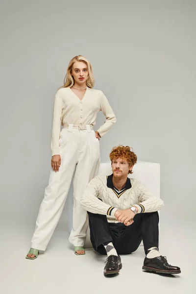 Trendy blonde woman looking at camera near white cube and redhead man sitting on grey, fashion — Stock Photo