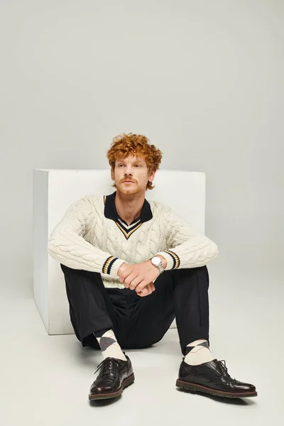 Stylish dreamy man in casual attire looking away sitting near white cube on grey, fashion concept — Stock Photo