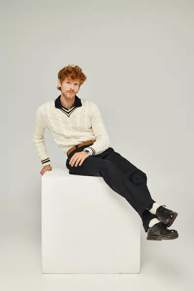 Trendy redhead man in casual attire sitting on white cube and looking at camera on grey backdrop — Stock Photo