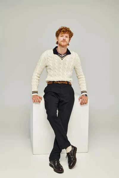 Fashionable redhead man in casual clothes sitting on white cube and looking at camera on grey — Stock Photo