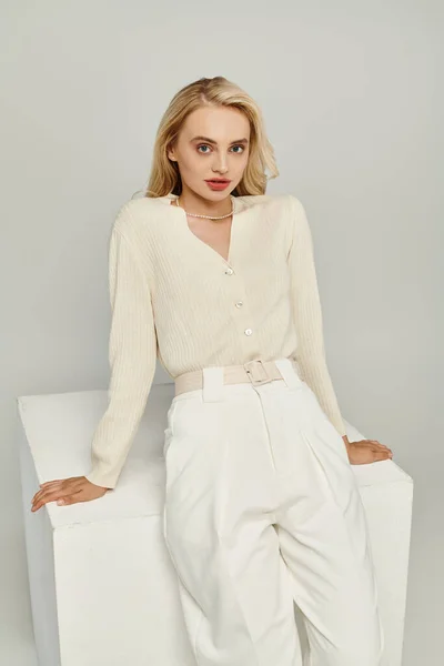 Aristocratic blonde woman in light clothes sitting on white cube and looking at camera on grey — Stock Photo