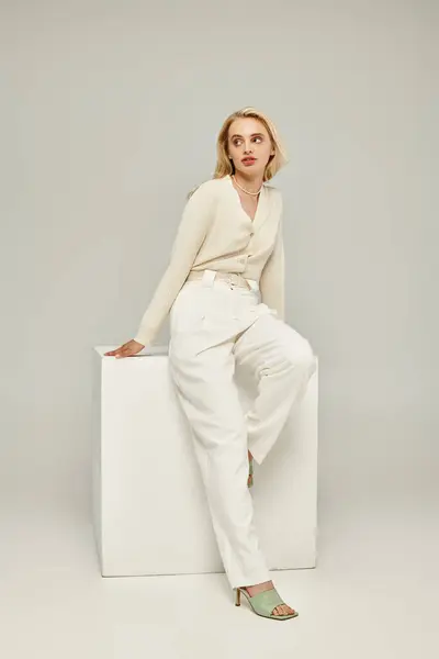 Fashionable blonde woman in light clothes sitting on white cube and looking away on grey backdrop — Stock Photo