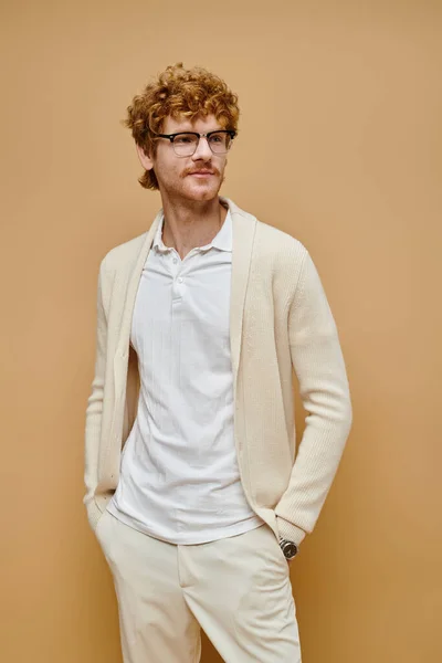 Homme rousse élégant souriant dans des lunettes détournant les yeux tout en se tenant avec les mains dans les poches sur beige — Photo de stock